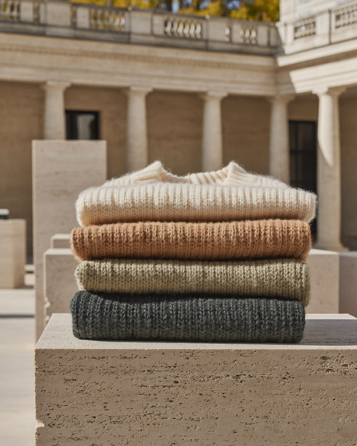 Gefaltete Strickteile in Alabaster, Salbeigrau und Tannengrün auf einer hellen Kalksteinplatte